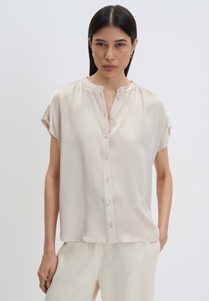 Frau mit langen dunklen Haaren trägt eine lockere, kurzärmelige, cremefarbene Bluse mit Knopfleiste und passende Hose, steht vor einem schlichten Hintergrund.