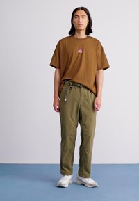 Bărbat cu păr lung, de culoare închisă, poartă un tricou maro supradimensionat, pantaloni verzi măslinii cu o curea și adidași albi, stând pe un fundal simplu.