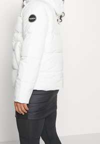 Vit vadderad jacka med huva, med en svart cirkulär logotyp på axeln. Texturerad, quiltad design med elastiska muddar.