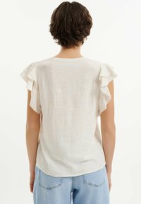 Blusa bianca di cotone con maniche a sbuffo, caratterizzata da dettagli floreali ricamati e una vestibilità leggermente ampia. La vista posteriore mostra un orlo curvo.