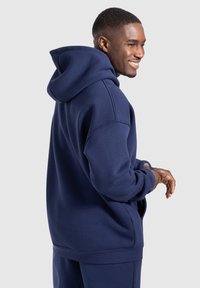 Felpa blu navy realizzata in morbido misto cotone. Presenta un grande cappuccio, spalle scese e polsini e orlo a costine. Texture liscia, vestibilità comoda.