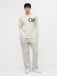 Jeune homme debout portant un pull crème avec le texte "Off", un pantalon beige clair à cordon et des baskets blanches sur un fond blanc uni.