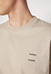 Beige bomull t-skjorte med rund hals, med to svarte broderte "SAMSONE" logoer på brystet. Glatt tekstur og avslappet design.