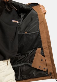 Person trägt schwarzen Oberteil und helle Hose, hält eine offene braune Jacke, die ihre schwarze Innenfütterung und Netzinnentasche zeigt.