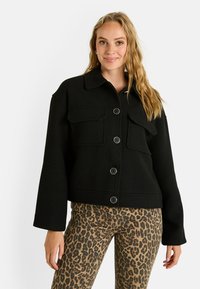 Veste noire courte avec un col, deux poches avant et des boutons ronds. Portée sur un pantalon à imprimé léopard. Texture douce, coupe ajustée.