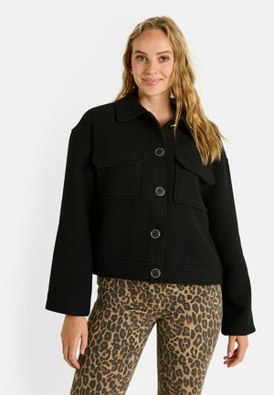 Chaqueta negra cortada con cuello, dos bolsillos frontales y botones circulares. Usada sobre pantalones de estampado de leopardo. Textura suave, ajuste entallado.