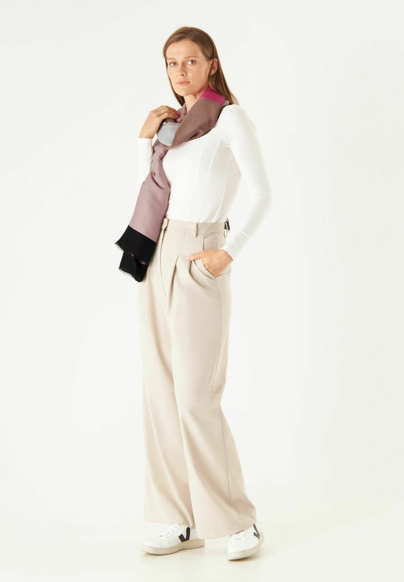 Lichte, multicolore sjaal hangt over een figuur in een strak wit topje en ruime, beige broek, gecombineerd met witte sneakers.