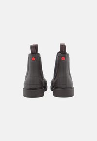 Καφέ καουτσούκ μποτάκια μέχρι τον αστράγαλο με υφή στις πλευρές, που διαθέτουν ένα κόκκινο κουμπί λογότυπου και λαβές. Λείος φινίρισμα, στρογγυλή μύτη, χοντρές σόλες.