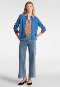 Cardigan blu con bottoni davanti, indossato sopra una camicia marrone a fantasia. Jeans larghi blu chiaro e scarpe con tacco marroni a slingback.