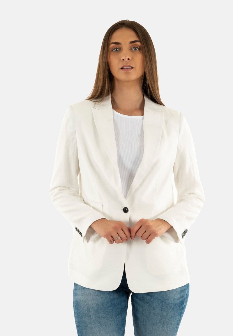 Mujer con cabello largo que lleva puesto un blazer blanco, una blusa blanca y jeans azules, ajustándose el blazer con ambas manos.