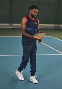 Mornarsko športno majico brez rokavov z rdečimi dodatki in modrimi črtami, kombinirano s mornarskimi trenirkami z modrimi črtami in belimi supergami, ki drži teniški rekvizit.