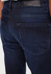 Tamno plave traperice imaju uski kroj s glatkom teksturom, dva stražnja džepa i kožnu oznaku s brendom na struku.