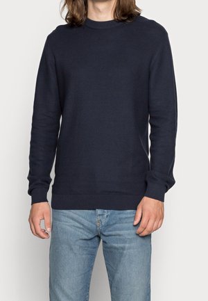 Mann trägt einen dunkelblauen Pullover mit langen Ärmeln und hellblaue Jeans vor einem schlichten, hellen Hintergrund.