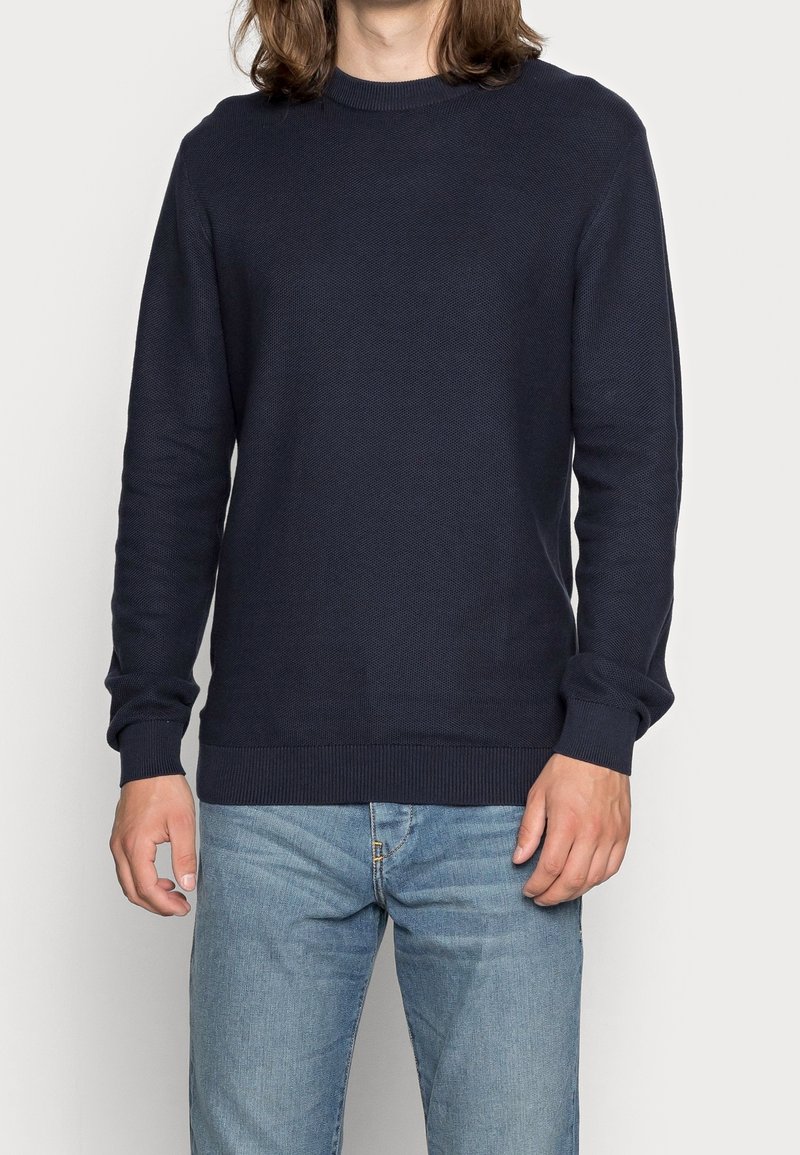 Homme portant un pull à manches longues bleu marine et un jean bleu clair sur un fond clair uni.