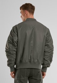 Olíva zöld bomber dzseki sima, fényes felülettel, bordás gallérral és mandzsettákkal, valamint gyűrt vállvarrással. Az alján logó hímzés található.