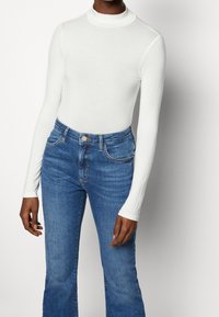 Vit långärmad polotröja bodysuit i kombination med blå högmidjade utsvängda jeans, som framhäver mjukt tyg och en figursydd silhuett.