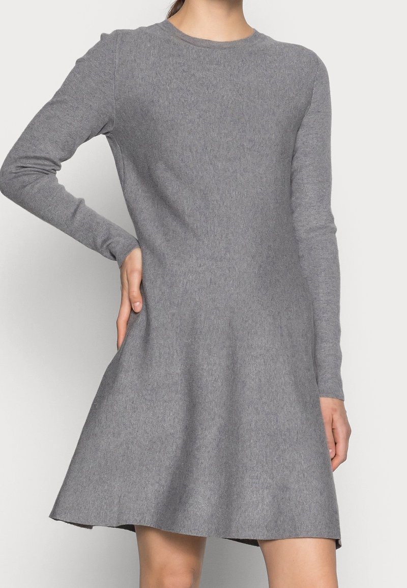 Femme portant une robe en maille grise à manches longues, longueur genou, avec un col rond, debout, une main sur la hanche.