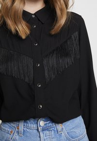 Blouse noire à fermeture boutonnée sur le devant, avec des détails à franges sur la poitrine. Manches longues et col classique. Portée avec un short en denim.