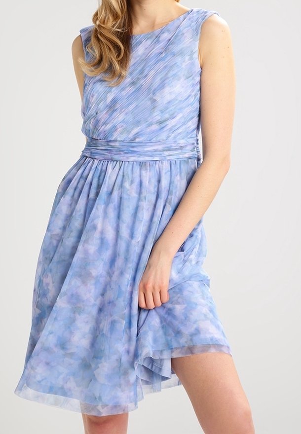 Femme portant une robe sans manches bleu clair à fleurs avec un corsage plissé et une jupe froncée, soulevant légèrement un côté avec la main.