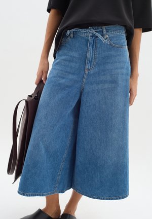 Kobieta nosząca szerokie, skrócone niebieskie jeansy, w połączeniu z czarnymi butami typu slip-on i trzymająca ciemnobrązową torbę typu tote.