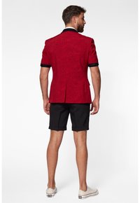 Crvena blejzerica s cvjetnim uzorkom, kratkih rukava, s crnim detaljima i otvorom na leđima, uparena s crnim šortsevima i bijelim tenisicama.