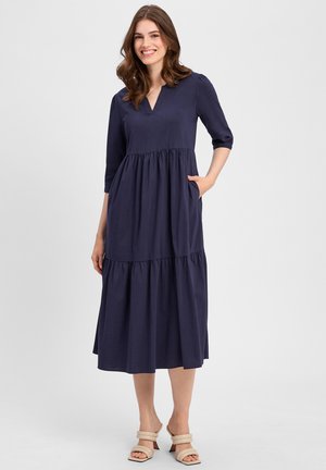 Navyblaue Baumwollkleid mit V-Ausschnitt, drei Viertelärmeln, gestuftem Design und Seitentaschen, kombiniert mit beigefarbenen Sandalen mit Riemchen.