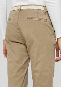 Beige chino's met achterste paspelzakken en een gevlochten crèmekleurige riem, gedragen met een wit langmouwen shirt, weergegeven vanaf de taille naar beneden.