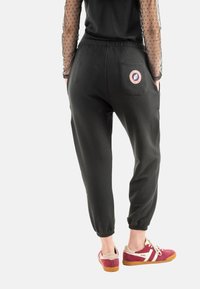 Pantalones de chándal negros con puños elásticos, con un bolsillo trasero que tiene un logo circular. Combinados con zapatillas de deporte color burdeos. Textura de tela suave.