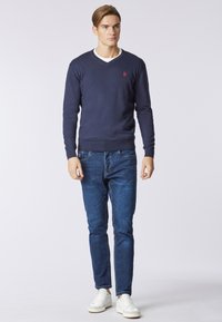 Maglione a v blu navy con un logo rosso sul petto, abbinato a jeans slim-fit blu e sneakers bianche. Texture liscia in tutto.