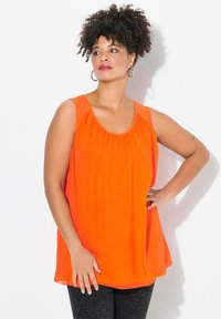 Top senza maniche di un vivace colore arancione con scollatura rotonda, dettagli arricciati sul busto e un tessuto leggero e fluente. Indossato con leggings neri a fantasia.