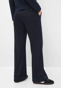 Personne portant un pantalon large bleu marine avec des ourlets retroussés et des baskets noires, se tenant avec une main dans la poche contre un fond blanc.