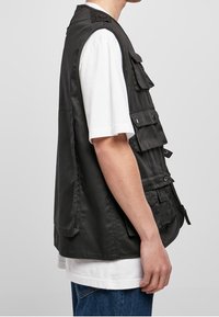 Musta käyttöliivi, valmistettu kevyestä kankaasta, jossa on useita taskuja ja rento istuvuus, pidetty valkoisen paidan päällä.