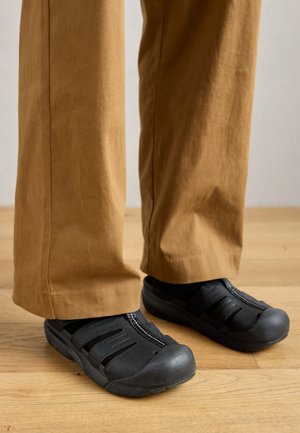 Scarpe nere senza lacci con suola in gomma, dotate di chiusura con zip e disegni a intaglio, abbinate a pantaloni larghi beige su un pavimento in legno.