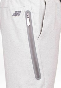 Hellgraue Jogginghose mit glatter Textur, ausgestattet mit einer Reißverschlusstasche und einer grauen Akzentlinie entlang des Reißverschlusses. Mit Logo auf dem Bein gestickt.