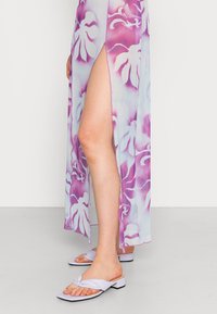 Vestido maxi floral em azul claro e roxo, com uma fenda alta. Usado com sandálias lilás de deslizar, com um design de tiras torcidas.