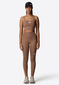 Sujetador deportivo marrón sin costuras y leggings de tiro alto a juego, ambos con un pequeño logo blanco. El modelo lleva puesto una gorra negra y zapatillas.