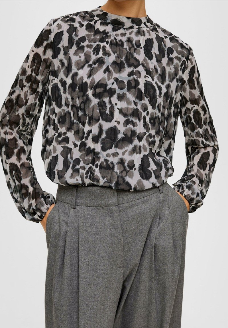 Femme portant un chemisier noir et gris à manches longues à imprimé léopard, rentré dans un pantalon gris plissé taille haute, les mains dans les poches.