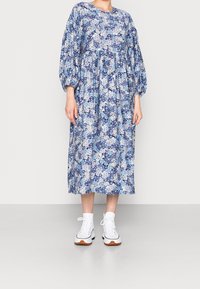 Žena v modro-bílé květinové midi šaty s dlouhými vyklenutými rukávy a bílými vysokými teniskami, stojící před jednobarevným pozadím.