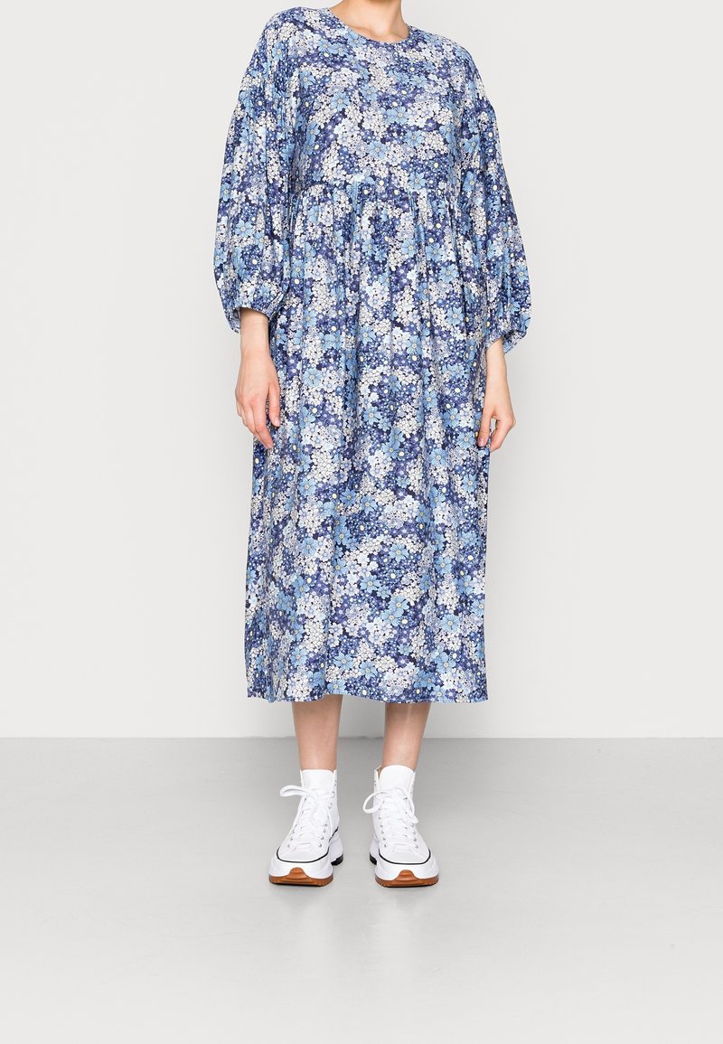 Žena v modro-bílé květinové midi šaty s dlouhými vyklenutými rukávy a bílými vysokými teniskami, stojící před jednobarevným pozadím.
