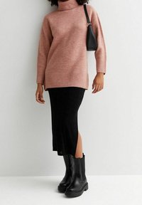 Maglione a collo alto rosa lavorato a maglia, gonna nera di lunghezza media e stivali neri con tacchi robusti. L'outfit ha una vestibilità rilassata e trame sovrapposte.