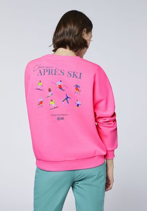 Person with short brown hair wears a bright pink sweatshirt with colorful skiing figures and "APRÈS SKI" text on the back.