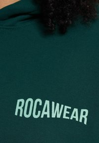 Dark green hoodie featuring a soft texture, with "ROCAWEAR" printed in light mint green. Includes a drawstring hood and ribbed cuffs.