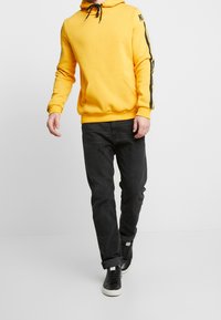 Felpa gialla con cappuccio e logo sulla manica, abbinata a jeans scuri e sneakers nere. Il design è casual e funzionale.