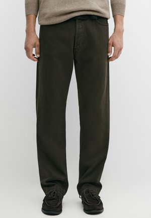 Man draagt losse zwarte broek, bruine trui en zwarte suède loafers, staand tegen een effen achtergrond.