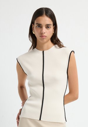Woman with long dark hair wears sleeveless, ribbed cream top with vertical black trim and gold hoop earrings, standing against white background.