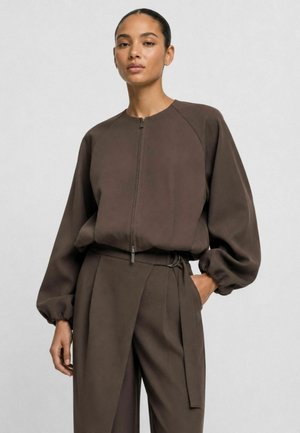 Vrouw draagt een bruine blouse met rits aan de voorkant en ballonmouwen, en bijpassende hoge taille broek met lussen voor een riem en plooien.
