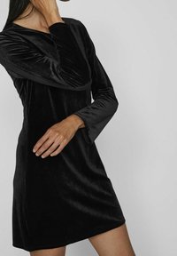 Vestido de terciopelo negro con mangas largas, escote en V y un corte relajado. La textura suave y el brillo liso acentúan el diseño general.
