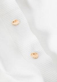 Tissu blanc à texture côtelée avec deux boutons arrondis de couleur dorée comme accents remarquables. Surface lisse et apparence douce.