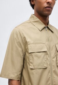 Άνδρας που φοράει μπεζ πουκάμισο με κοντά μανίκια, δύο μπροστινές τσέπες με καπάκι και φερμουάρ στο μπροστινό μέρος, εμφανιζόμενος από τους ώμους μέχρι το μέσο του κορμού.