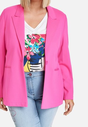 Frau trägt einen knallpinken Blazer über einem weißen Top mit buntem Blumenmuster und hellblauen Jeans.
